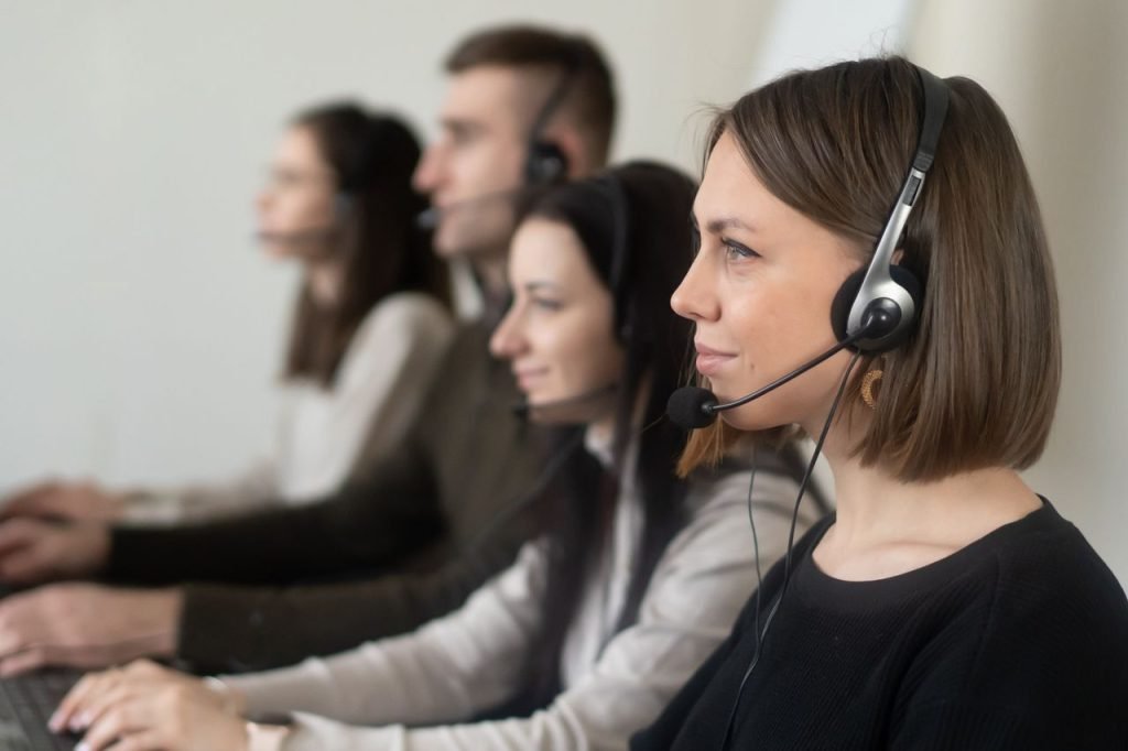 Side view of young coworkers in casual clothes sitting in light workplace with headphones with microphone while surfing on laptop